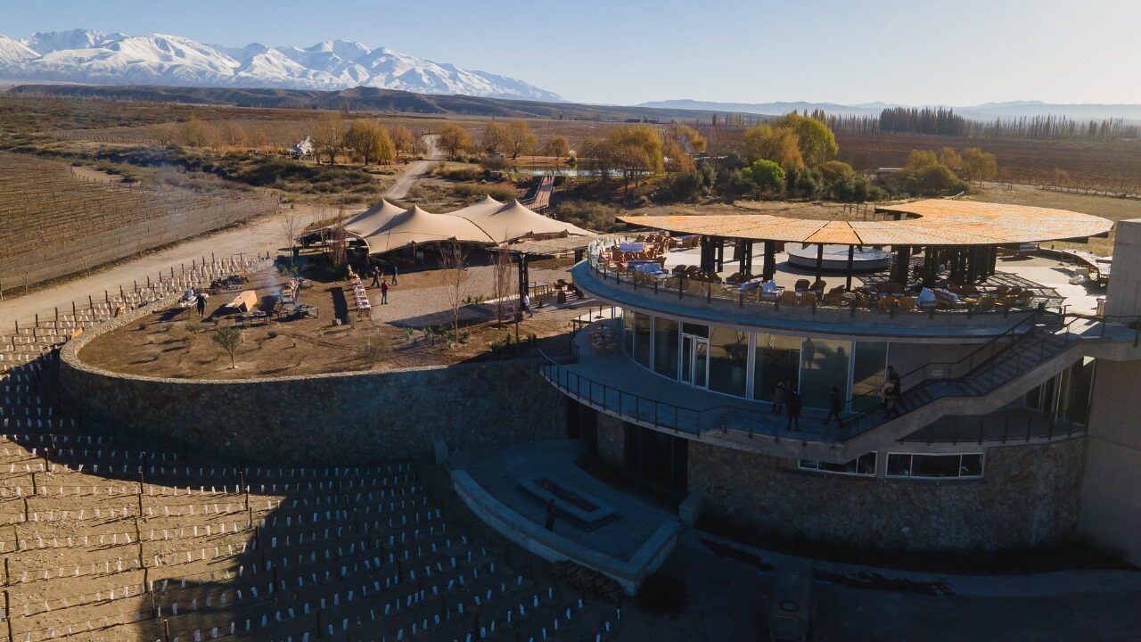 Vista aérea de uma bodega circular cercada de vinhedos, com tenda montada ao lado e os Andes ao fundo
