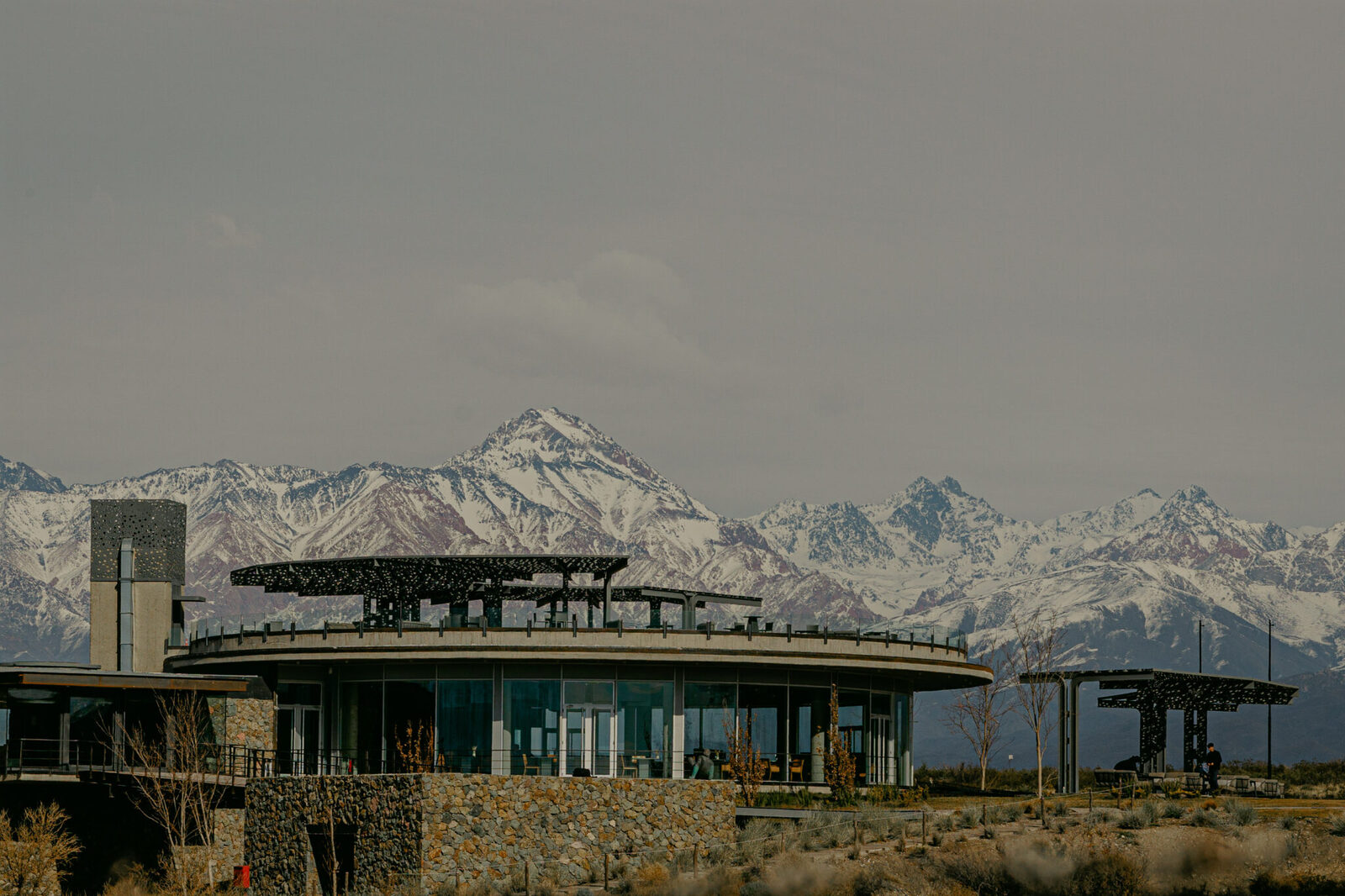 Bodega circular de pedra e vidro com pérgolas vazadas, e a Cordilheira dos Andes nevada atrás, sob luz cinzenta de inverno