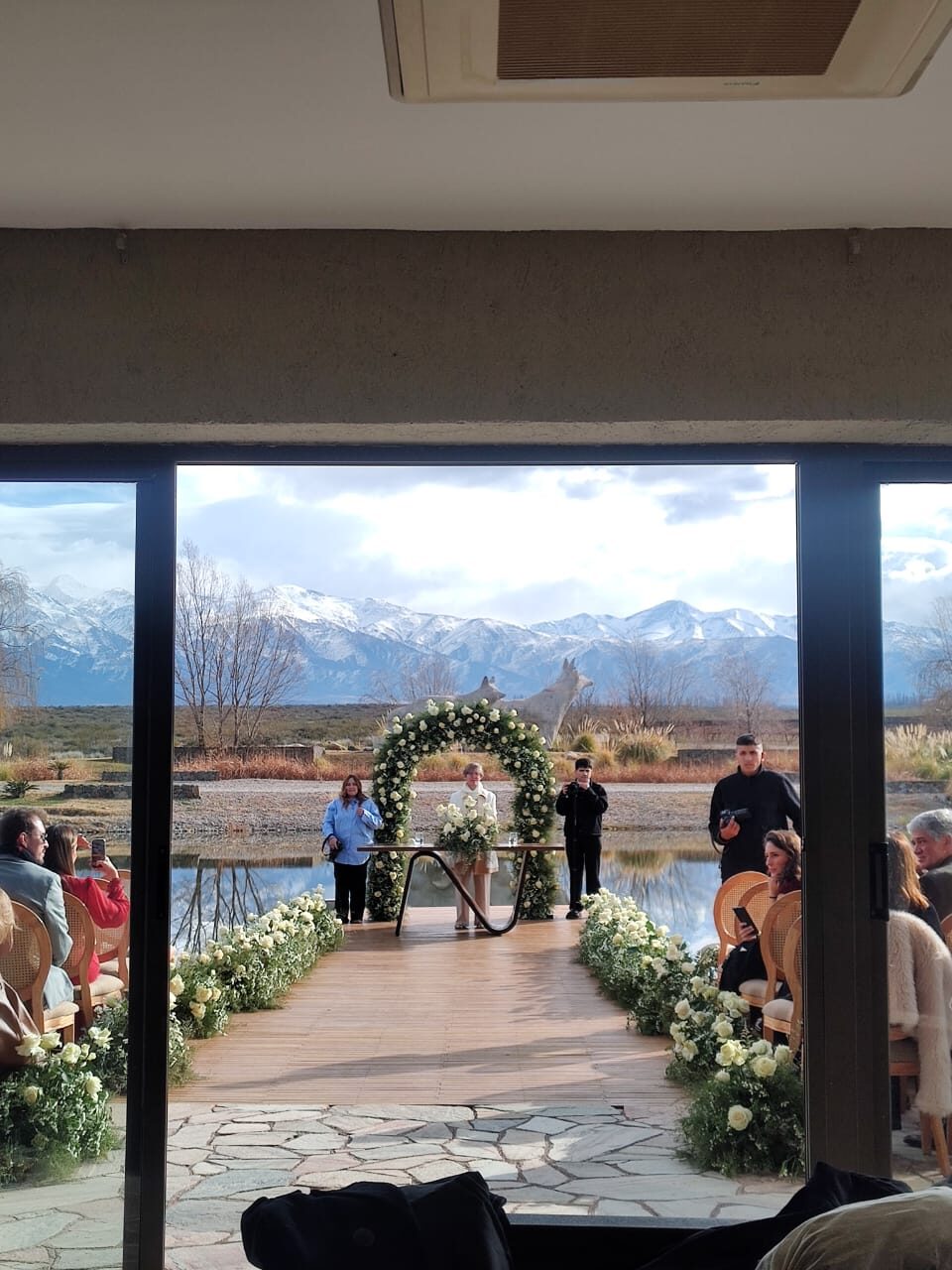 Cerimônia de casamento com arco de flores brancas diante de um espelho d'água, convidados sentados e os Andes nevados ao fundo