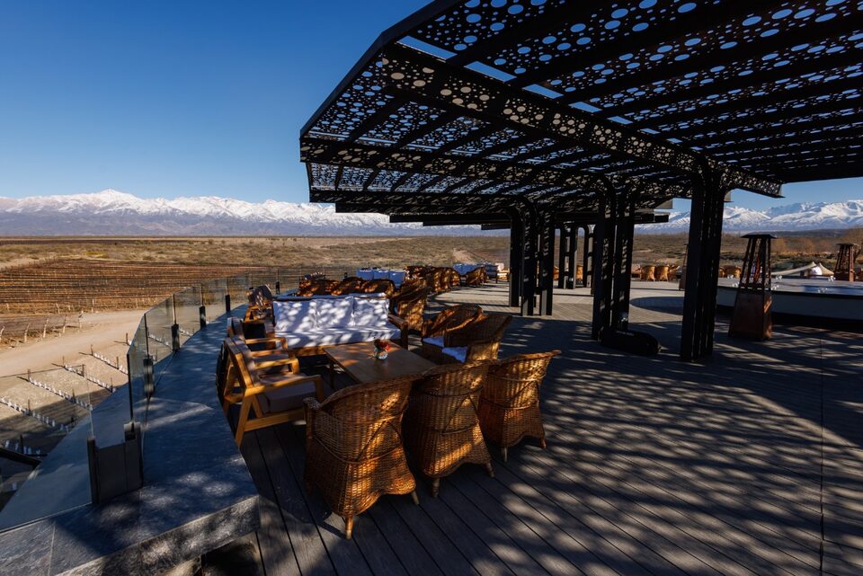 Mesa comprida posta sob pérgola de ferro no terraço da bodega, com os Andes nevados ao fundo